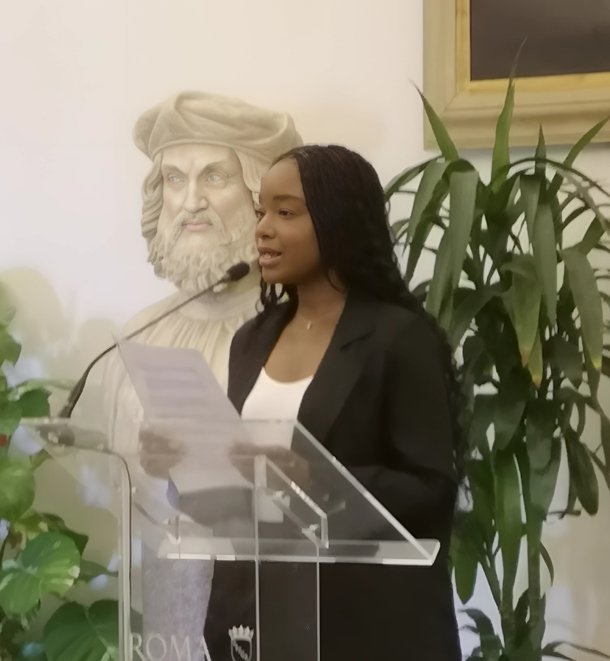 Donna parla al podio accanto a busto classico.