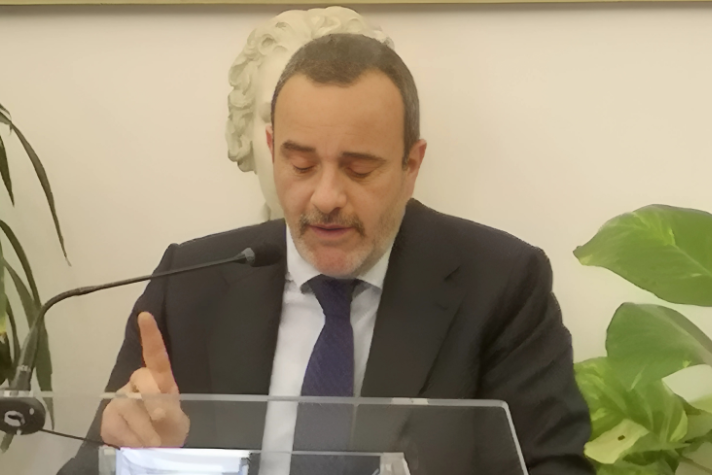 Uomo in abito tiene discorso dietro podio trasparente.